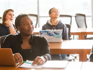 Students in a classroom, the student in the foreground looks at speaker thoughtfully.