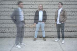 Three people stand on a street, but their images are partly transparent against the brick wall behind them