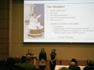 Three presenters stand in front of a screen displaying the WeeBot