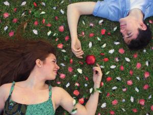 Actors from 'The Diary of Adam and Eve' lay on grass peering at each other. Flower pedals cover the ground and an apple is between their hands.
