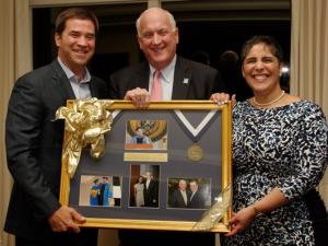 Two men and a woman pose as they hold a framed piece that showcases several photos and a medal. 