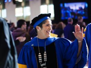 A woman in ceremonial academic cap and robes smiles and waves.