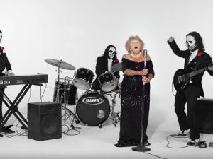 An older woman in black sings in front of a goth band