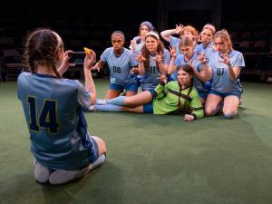 A group of female-identifying actors in blue soccer uniforms. They have orange slices in their mouths and are posing for a camera and making "wolf" expressions with their mouths and hands. Another actor is taking their picture with a cell phone.