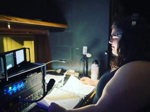 A stage manager wearing a headset in front of a calling script and video monitors.