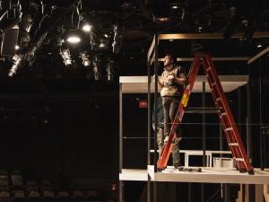 A TPD student stands on a ladder which sits on a platform. He is holding a drill and looking at lights.
