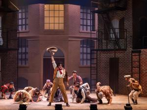Actors in Newsies. Actors are crouched on the ground around a main actor with his hand raised triumphantly in the air.