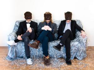 Three people with suits and bear masks sit on a couch covered in silver tinsel