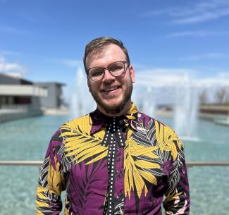 Dev is wearing a purple and yellow floral button up shirt while smiling in front of the Ithaca College fountain on a sunny day. 