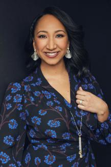 Photo of a smiling woman with medium brown skin and dark hair. She is wearing a black dress with blue flowers and large silver earrings