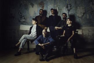 Six people sitting on chairs, on the floor, and standing against a wall