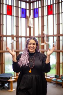 Lauren Kelly is seen wearing a black dress, brown mala beads, a ring on their necklack, lilac hair, smiling at the cameral with arms open and facing up. They are stanging in the Muller Chapel Gathering Hall with windows behind themme. 