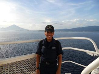 A woman in a Navy uniform stands on the deck of a Naval vessel.