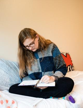 A student in a blue and gray checked sweater is seated on a bed writing in a notebook.