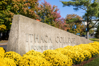 Ithaca College sign found at entrance to the college. The sign is a large cement block, with pale letters spelling out "Ithaca College, Founded 1892" on the side of it.  The sign is surrounded by yellow flowers, and there are colorful autumn trees in the background.