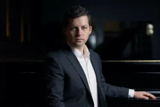 pianist Henry Kramer standing beside a piano, wearing a dark suit jacket and a white shirt
