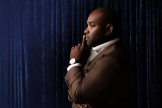 tenor Lawrence Brownlee stands in profile in front of a blue curtain, with his finger to his lips