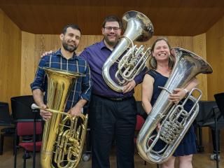 Three people stand on a stage holding tubas