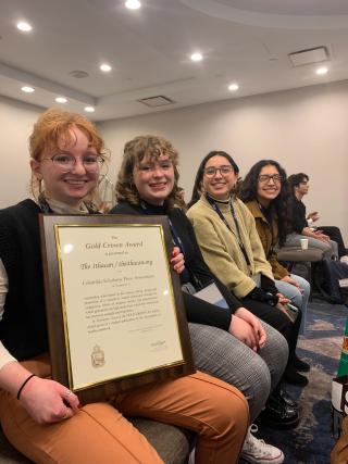 Four students are seated at a conference, with the one to the far left holding a CSPA Gold Crown Award.