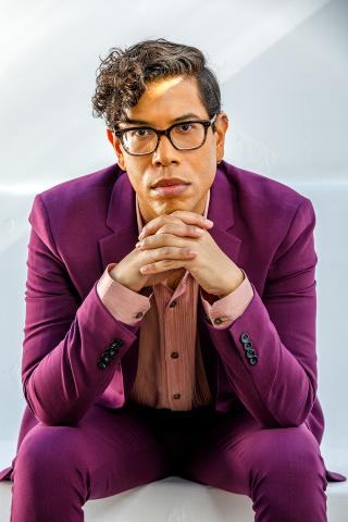 Image of Steven Canals posing for camera with white/grey background. Canals is wearing a magenta suit with pink textured dress shirt and glasses. He is leaning forward with his chin resting on his hands and elbows resting on top of his thighs.