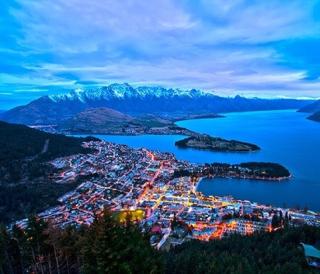 An aerial shot of the city of Christchurch, New Zealand at dusk.  Shows city lights, the water in the bay, an island, and snow-capped mountains in the distance. The sky is cloudy and the clouds reflect pink from the sunset.