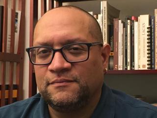 Carlos is posing for headshot in front of bookshelf. Carlos is wearing a blue shirt and glasses. Carlos has facial hair around chin. Carlos is looking at the camera with mouth closed.