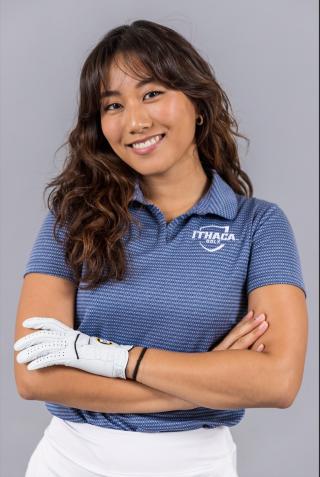 Cristea is posing for headshot with arms crossed in front on grey background. Is wearing light blue striped Ithaca Golf polo and white pants. Has a white golf glove on left hand. Has brown hair extending past shoulders.