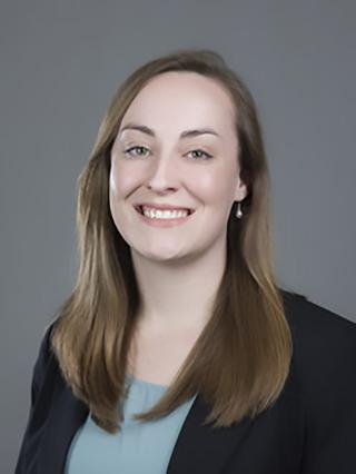 Headshot image of Sam on a grey background. Sam is wearing a light blue shirt and navy blue blazer. She is looking directly at the camera and smiling.