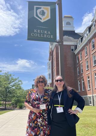 Mish Lenhart and Jess Shapiro in front of Keuka College flag and building