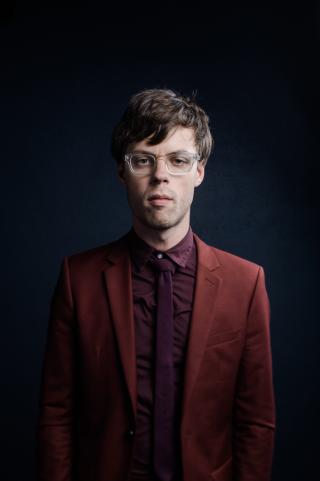 a person in a burgundy suit and glasses stands against a dark background