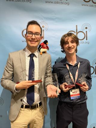 Two male college students stand with five awards in front of a CBI step and repeat.