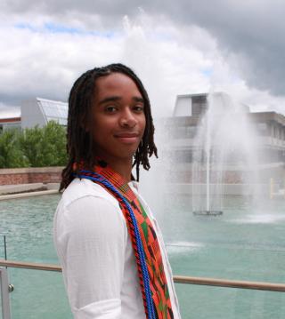 Image of Ty Benners posed in from of Dillingham Fountains. Ty is positioned side-facing looking right to left in the image. Ty is wearing a white shirt with a multi-colored stole.