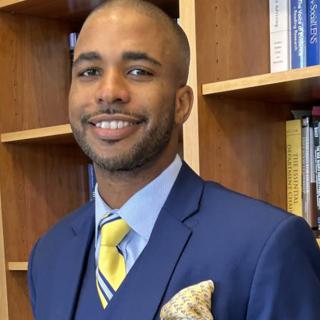 Stanley Bazile is posed for headshot in front of wooden bookshelves. He is wearing a blue suite with light blue shirt and yellow tie with blue stripe. He has a light yellow patterned pocket square. He is looking at the camera and smiling.