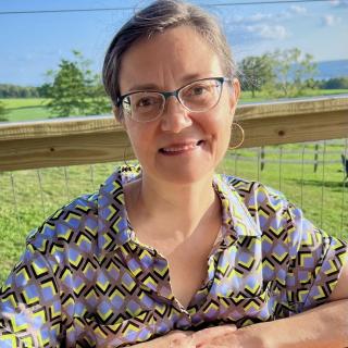 white female sitting hands together smiling at the camera. she has brown hair, glasses and is wearing a patterned shirt. 